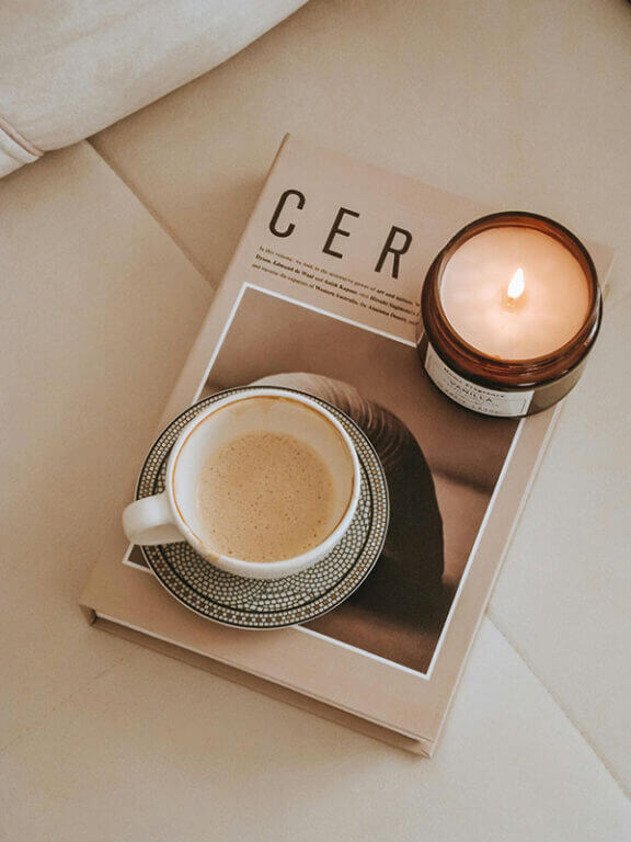 A cup of coffee on a saucer next to a lit candle, resting on an open book, all placed on a table.