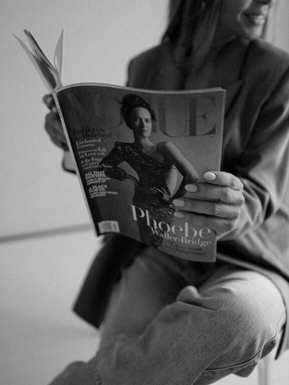 A woman with a black and white attire holding a magazine indoors, leaning against a wall.