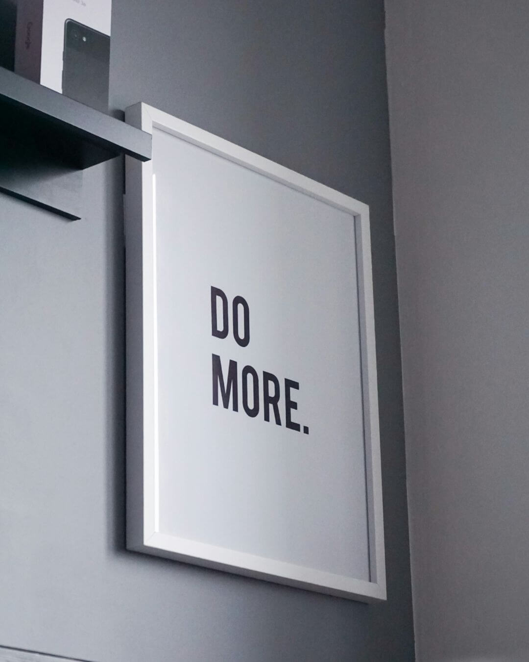 A white rectangular sign mounted on a wall that reads "DO MORE."