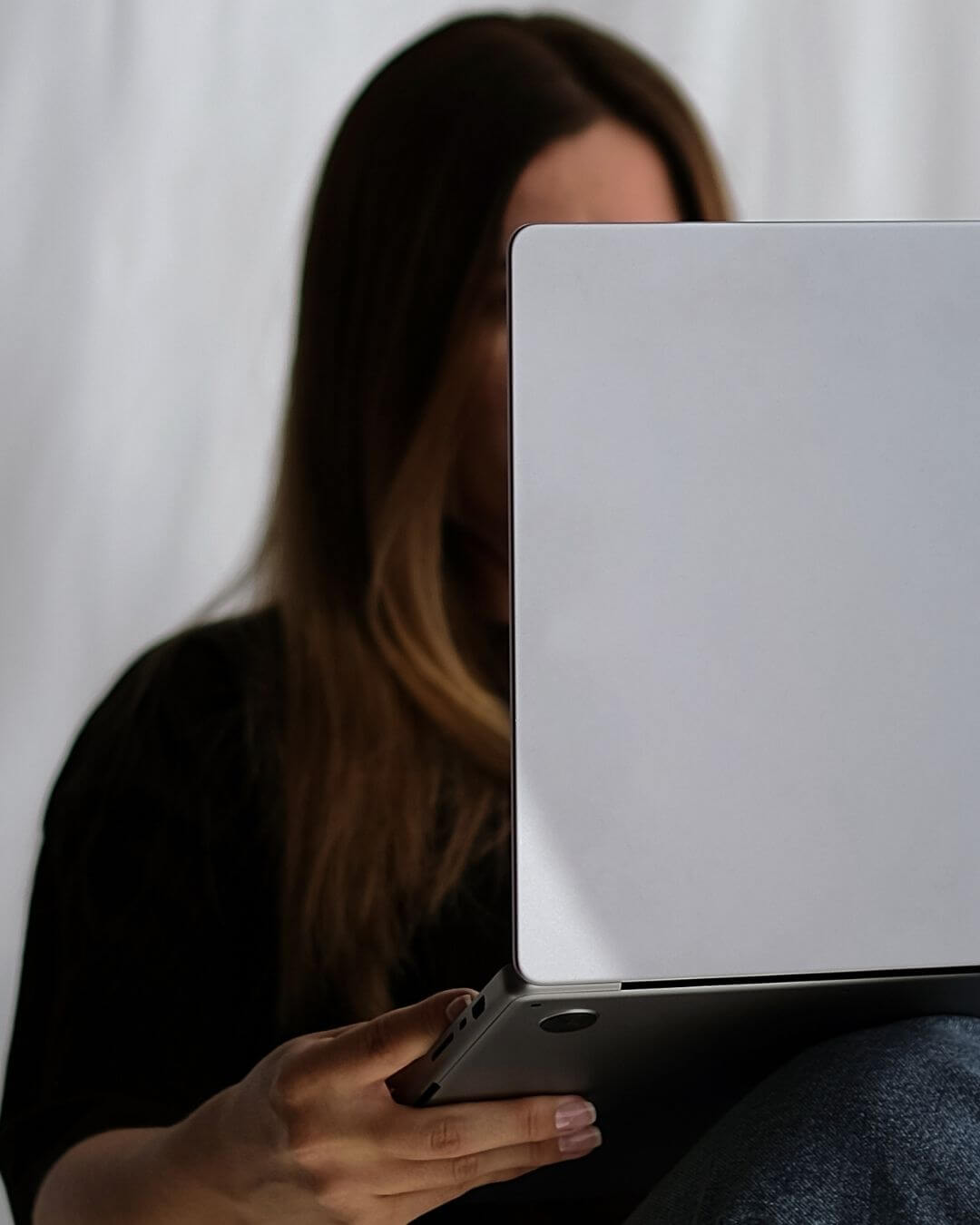 A woman sitting indoors, holding a laptop in her hands while engaging with the device.