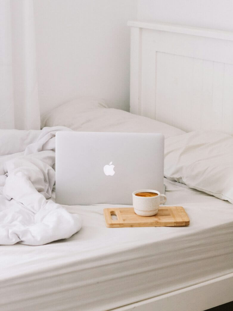 A laptop and a cup of coffee placed on a bed with white linens and pillows.